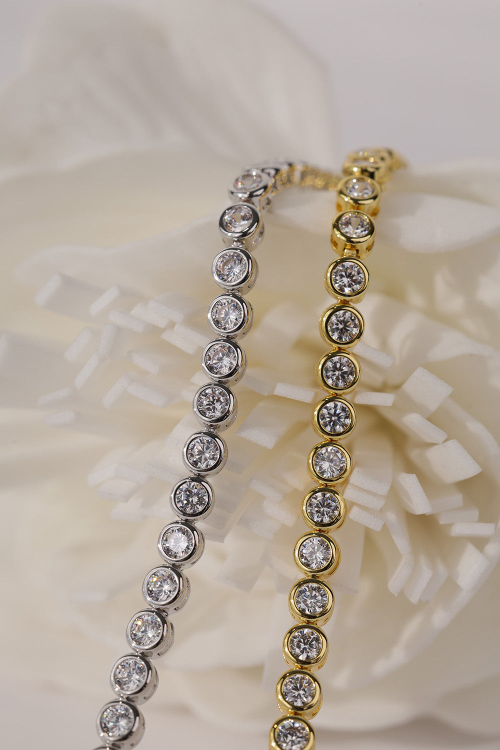 Two tennis bracelets with round clear stones, one silver and one gold, displayed on a soft white flower background.