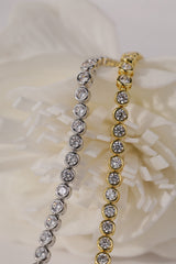 Two tennis bracelets with round clear stones, one silver and one gold, displayed on a soft white flower background.