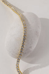 A gold tennis necklace with clear stones, draped elegantly over a smooth white stone on a light beige background.