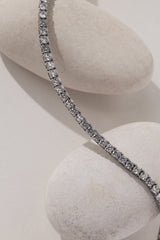 A silver tennis nceklace with clear stones, draped elegantly over two smooth, light-colored decorative stones against a neutral light gray background.