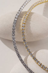 Two tennis-style necklaces draped over a light stone display form, one in silver-toned metal and one in yellow gold-toned metal, both set with clear stones.