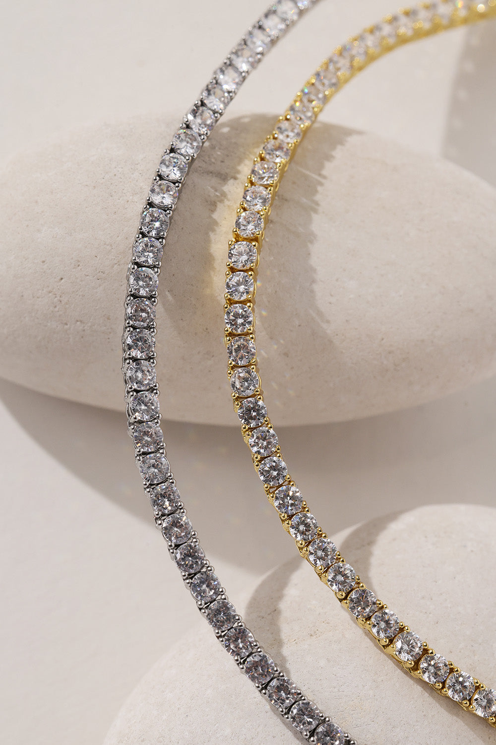 Two tennis necklaces draped over a light stone display form, one silver and one gold, both set with clear stones.