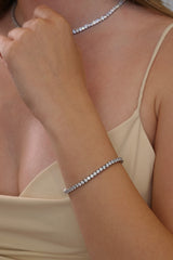 A close-up of a model wearing a delicate silver tennis bracelet on her wrist, with a matching silver choker necklace and a beige camisole top.