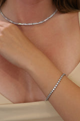 A close-up lifestyle shot of a model wearing a delicate silver tennis necklace and a matching silver tennis bracelet, styled with a cream-colored top.