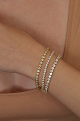 Three tennis-style bangle bracelets with clear stones, including two gold-toned and one silver-toned, stacked on a person's wrist.