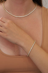 A close-up of a model wearing a delicate gold tennis necklace and a matching gold tennis 3mm bracelet, paired with a beige tank top.