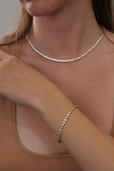A close-up of a woman wearing a delicate gold tennis necklace and a matching gold tennis bracelet, both adorned with small clear stones.