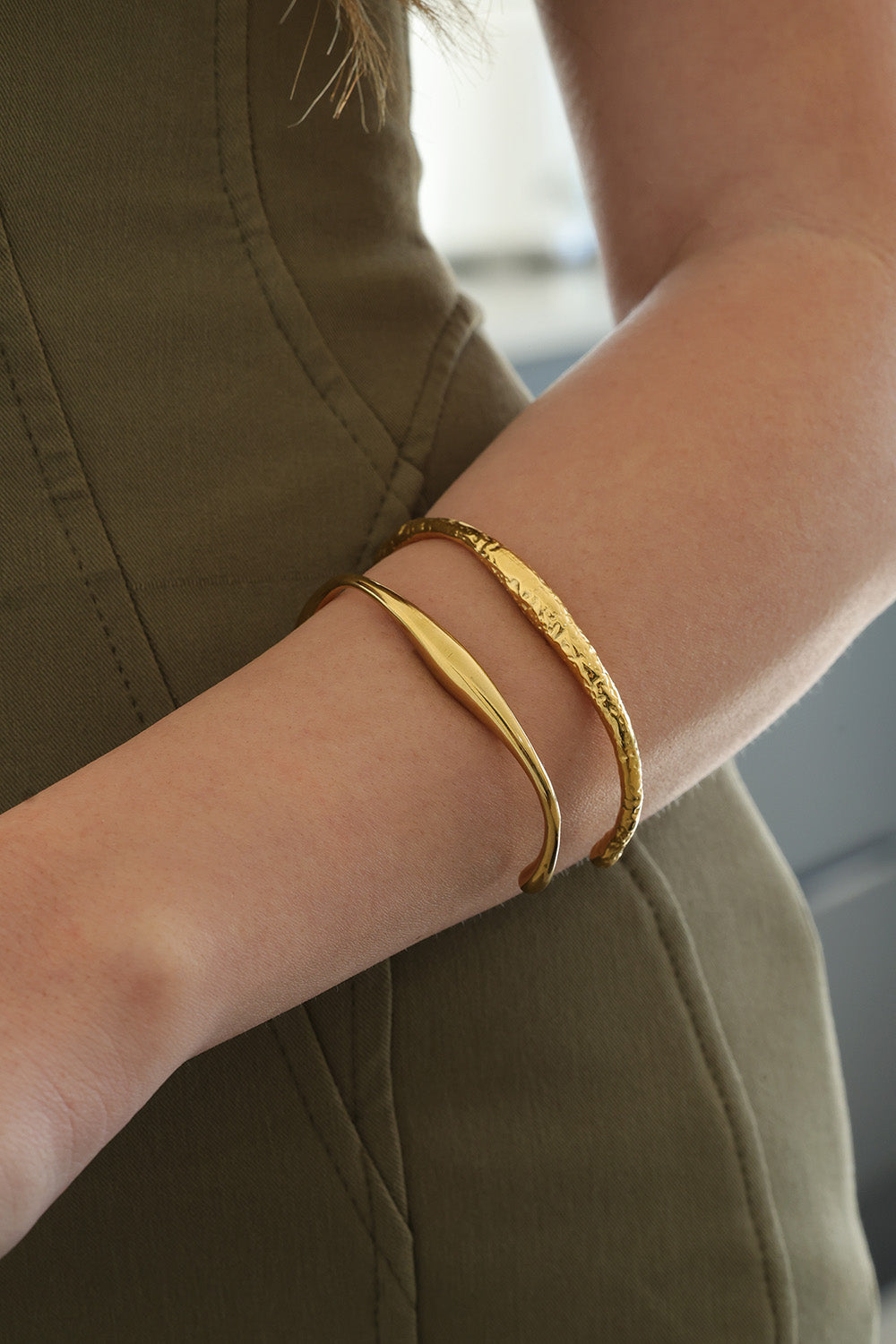 Two gold-toned bangle bracelets worn on a person's wrist, with one smooth curved bangle and one textured bangle, shown against an olive green top.