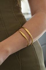 Two gold-toned bangle bracelets worn on a person's wrist, with one smooth curved bangle and one textured bangle, shown against an olive green top.