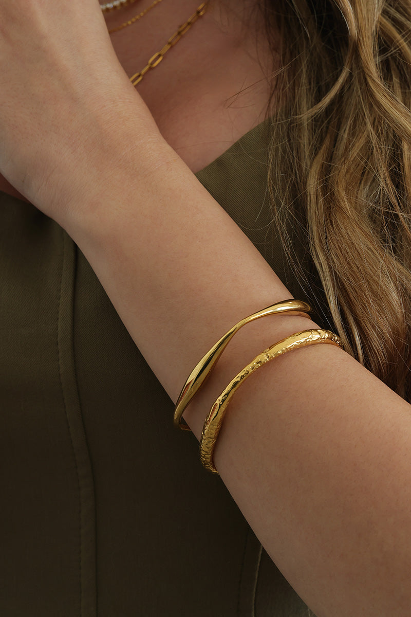 A close-up of a woman's wrist wearing three gold-toned bangles, including one textured bangle, against an olive green top.