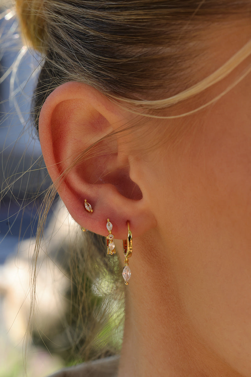 Close-up of a person's ear wearing multiple small gold earrings, including a huggie hoop with a clear teardrop crystal and a dangling gold charm with a clear stone.