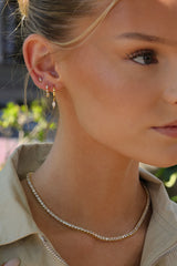A close-up lifestyle shot of a woman wearing a delicate gold tennis necklace and matching small gold hoop earrings with clear stones.