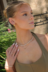 A woman wearing delicate gold jewelry, including a pearl necklace, hoop earrings, rings, and a bracelet, posing outdoors against a background of greenery and a stone wall.