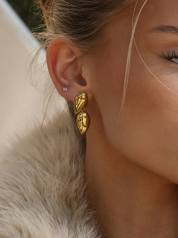 A close-up of a model's ear wearing a pair of chunky, textured gold drop earrings and a small silver stud earring, with her hair and a light-colored fur collar visible.