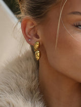 A close-up of a model's ear wearing a pair of chunky, textured gold drop earrings and a small silver stud earring, with her hair and a light-colored fur collar visible.