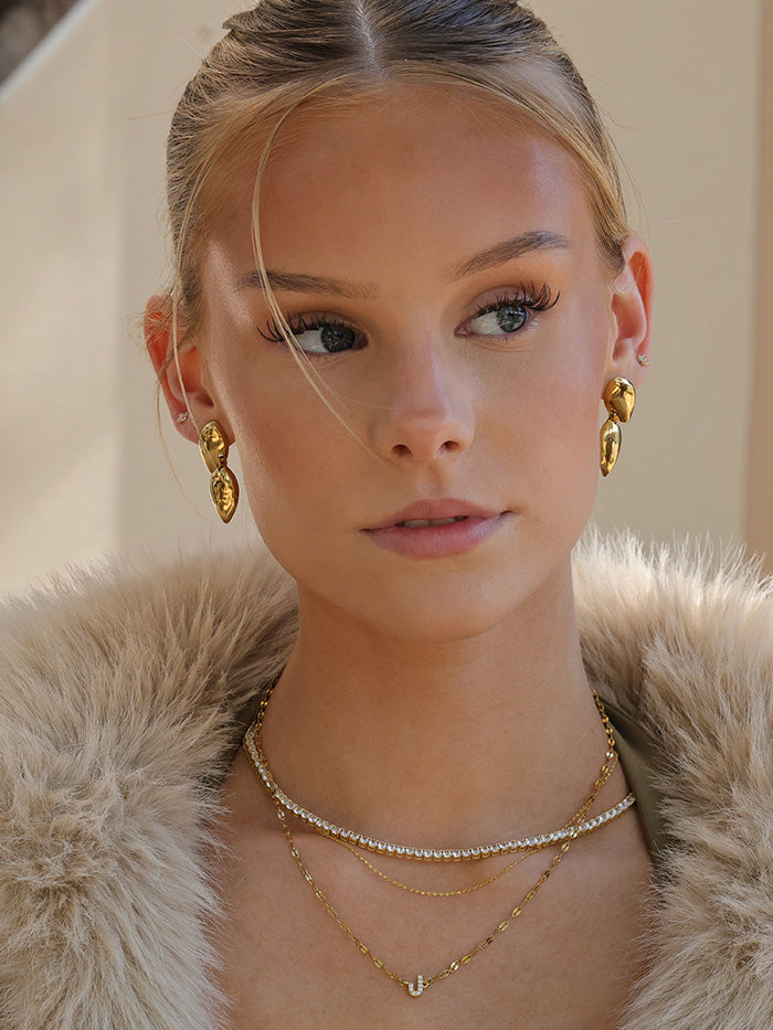 A close-up portrait of a woman wearing gold jewelry, including a gold teardrop earring and layered gold necklaces, paired with a beige faux fur collar.