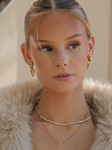 A close-up portrait of a woman wearing gold jewelry, including a gold teardrop earring and layered gold necklaces, paired with a beige faux fur collar.