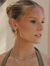 A close-up side profile of a woman with blonde hair in a bun, wearing a gold leaf-shaped hoop earring and layered gold necklaces, including a delicate diamond-studded chain.