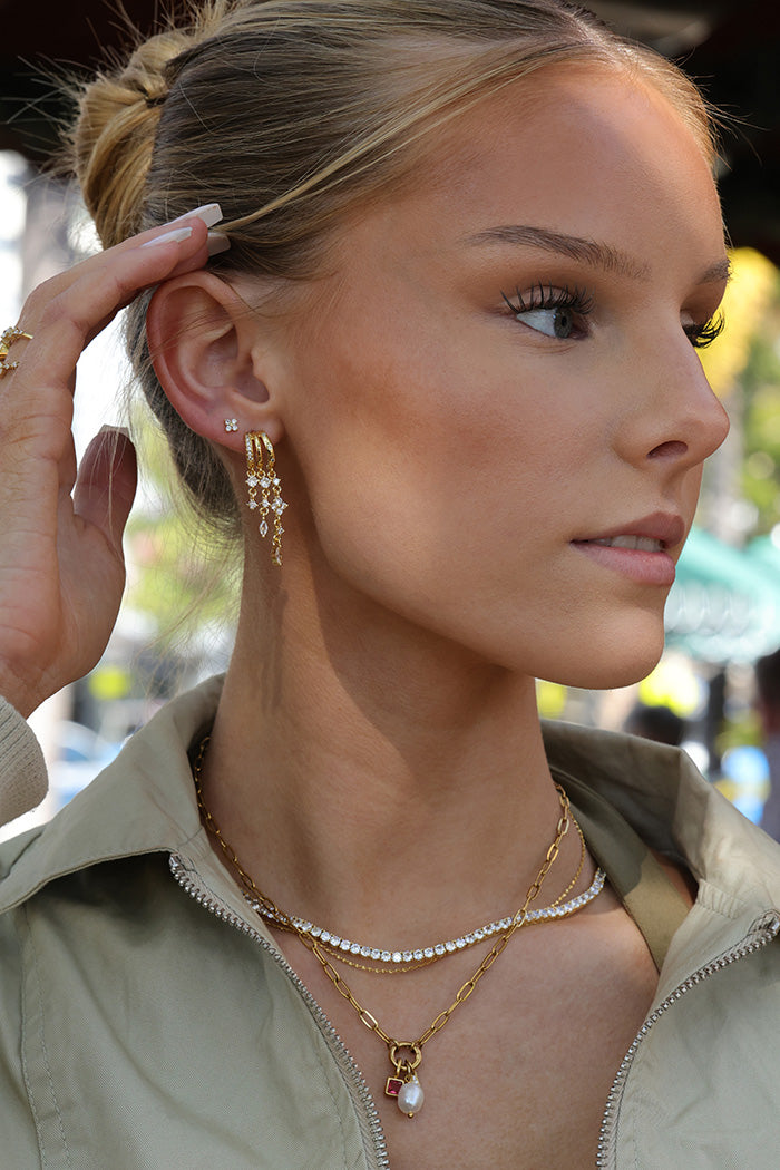 A close-up lifestyle shot of a woman wearing gold jewelry, featuring a gold dangle earring with clear crystals, layered gold necklaces, and a gold ring, paired with a beige zip-up jacket.