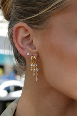 A close-up of a woman's ear wearing a gold huggie hoop earring adorned with clear crystals and a dangling cascading earring with gold chains and crystal drops.
