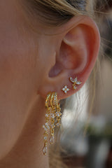 A close-up of a person's ear wearing a pair of gold hoop earrings with dangling floral and teardrop crystal charms.