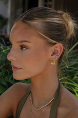 A close-up side profile of a woman with blonde hair in a bun, wearing gold huggie hoop earrings with small white crystal clusters and layered gold necklaces, set against a blurred green garden background.
