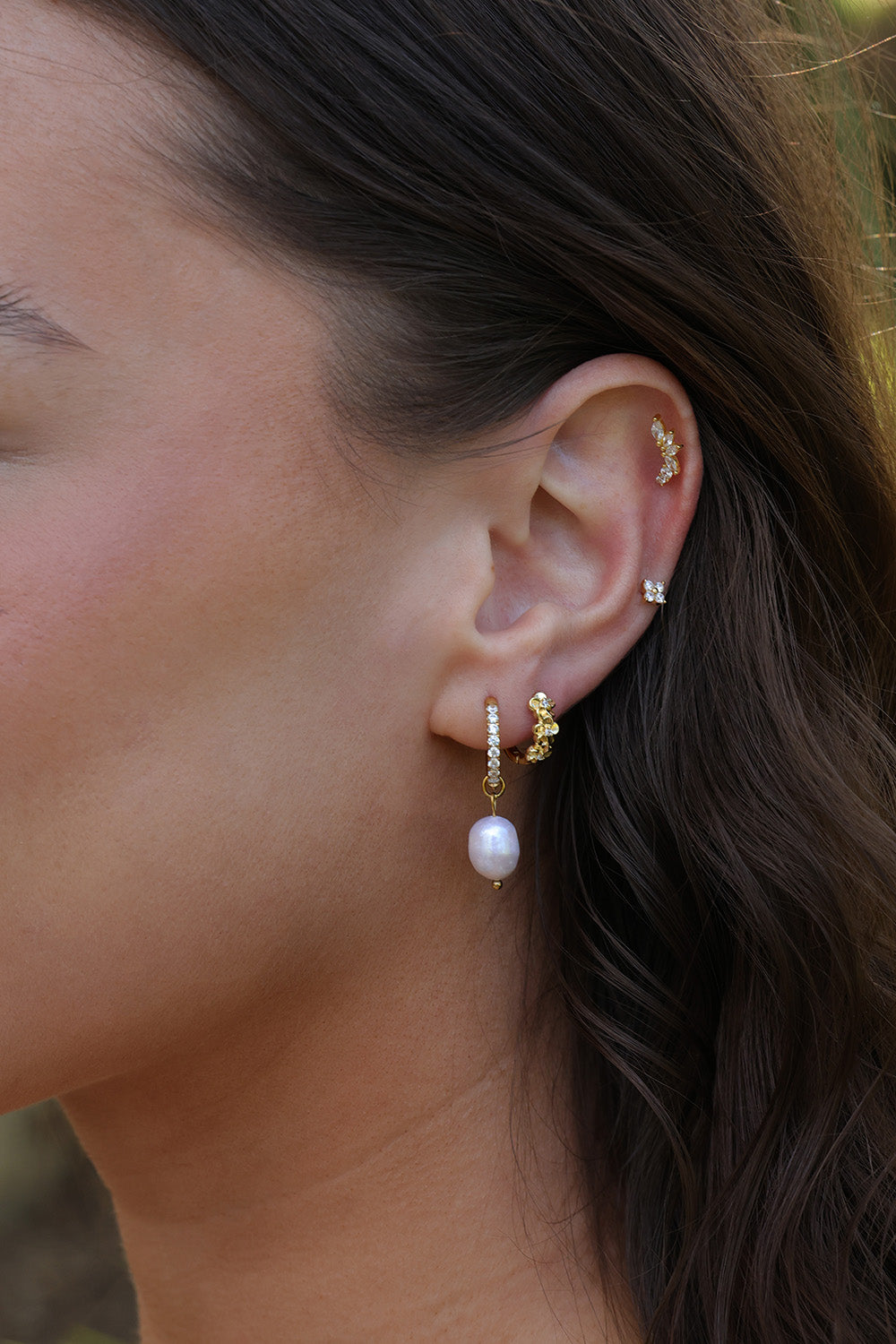 A close-up of a woman's ear wearing a gold huggie hoop earring with a dangling white pearl, along with small gold ear cuffs.