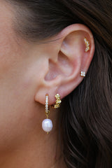 A woman's ear styled with a curated four-piece gold ear stack — a cubic zirconia huggie with baroque pearl drop, a gold floral huggie, a marquise climber and a clover stud.