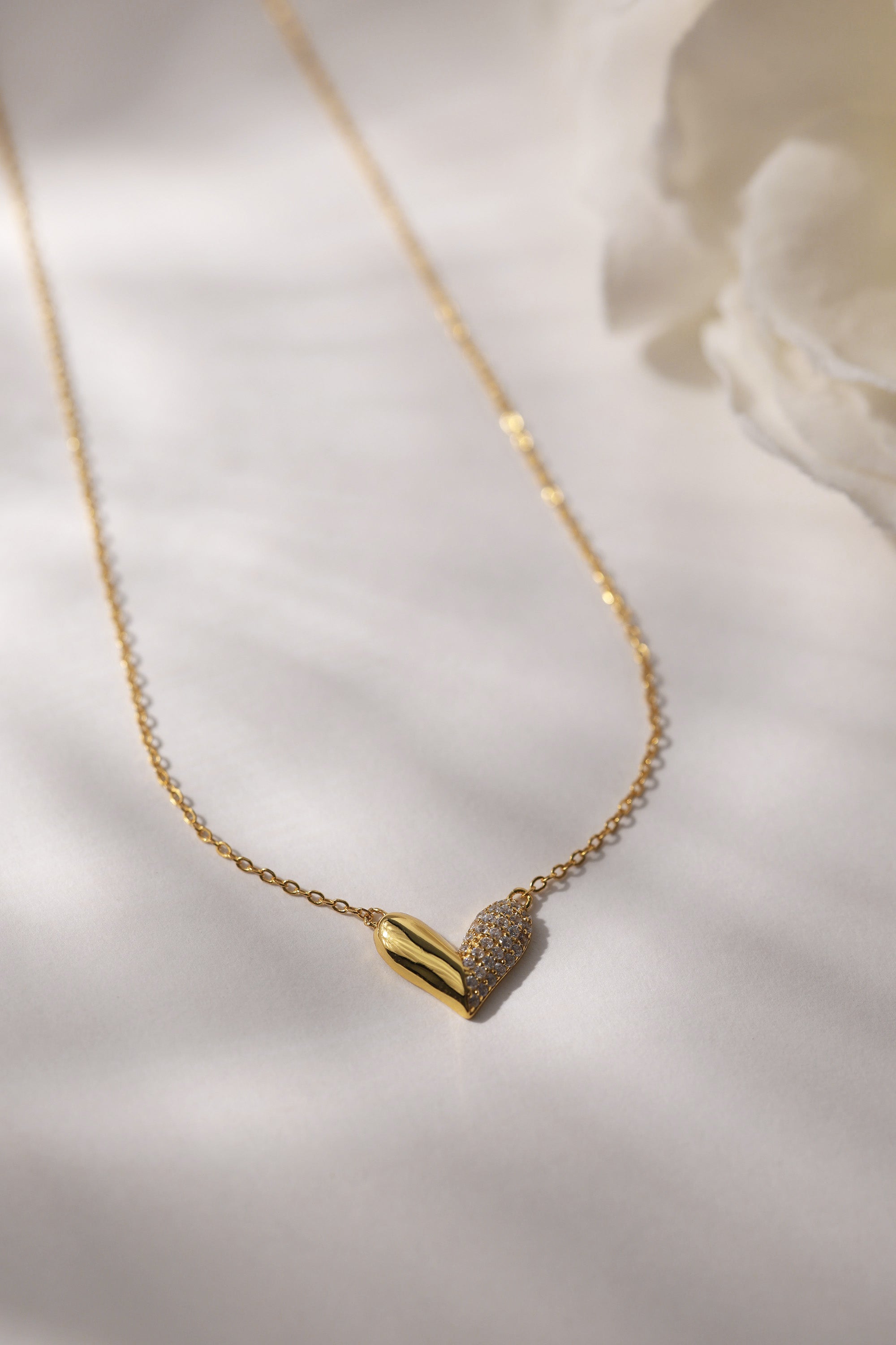 Elegant heart-shaped necklace in 18K gold-plated sterling silver with half pavé crystal detailing, displayed against a neutral background. Hypoallergenic and tarnish-resistant design.