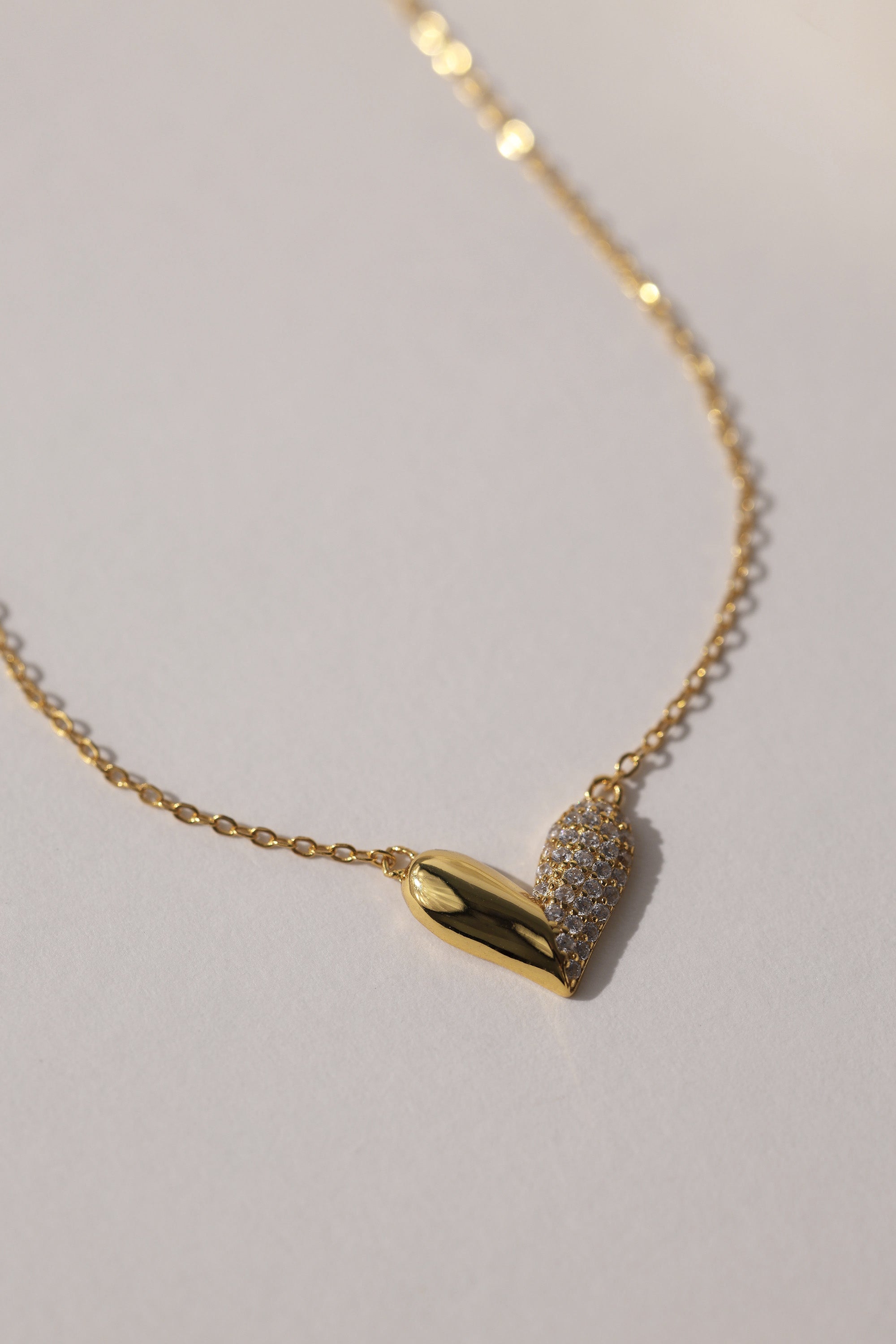Elegant heart-shaped necklace in 18K gold-plated sterling silver with half pavé crystal detailing, displayed against a neutral background. Hypoallergenic and tarnish-resistant design.