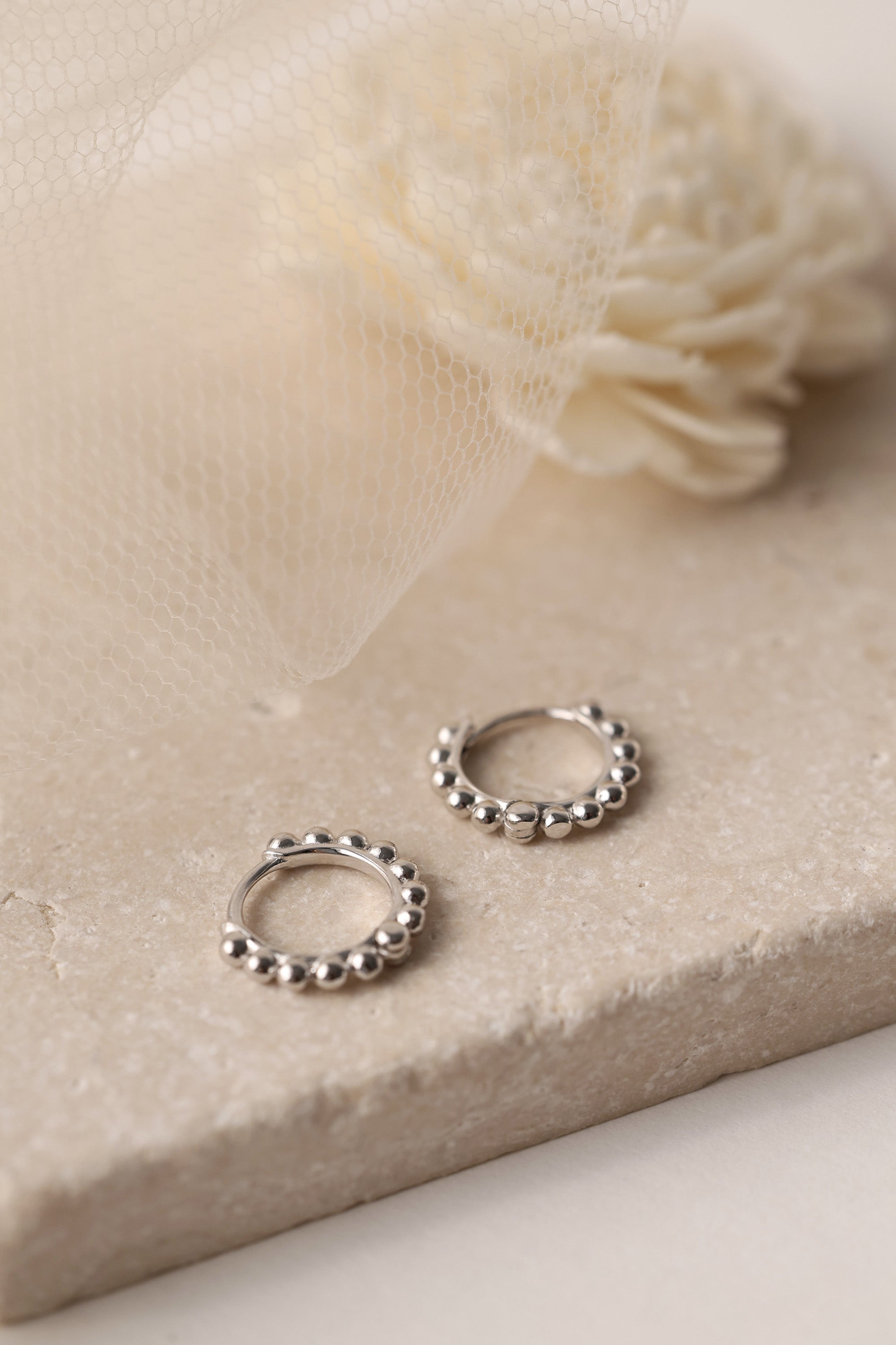 Sterling silver beaded hoop earrings in 8mm and 9mm sizes, featuring polished spherical detailing for a dainty, minimalist look. Displayed on a neutral textured surface with soft floral accents.