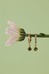 A pair of gold huggie hoop earrings with small clear stones and green peridot faceted drop charms, hanging from a green flower stem against a solid light green background.