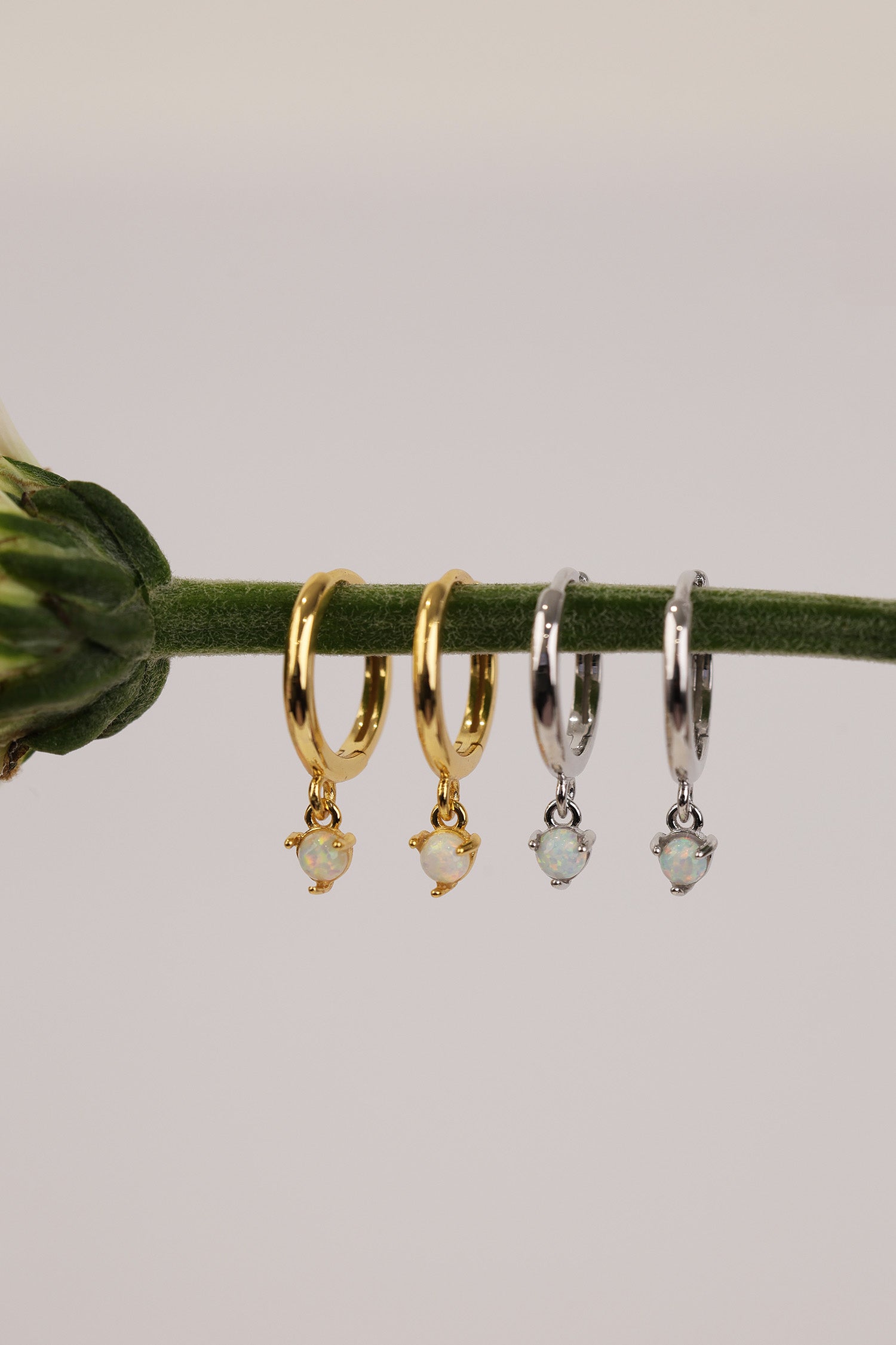 Four small hoop earrings hanging from a green stem against a light gray background. Two hoops are gold-toned with a tiny opal stone dangle, and two are silver-toned with matching pale stone dangles.