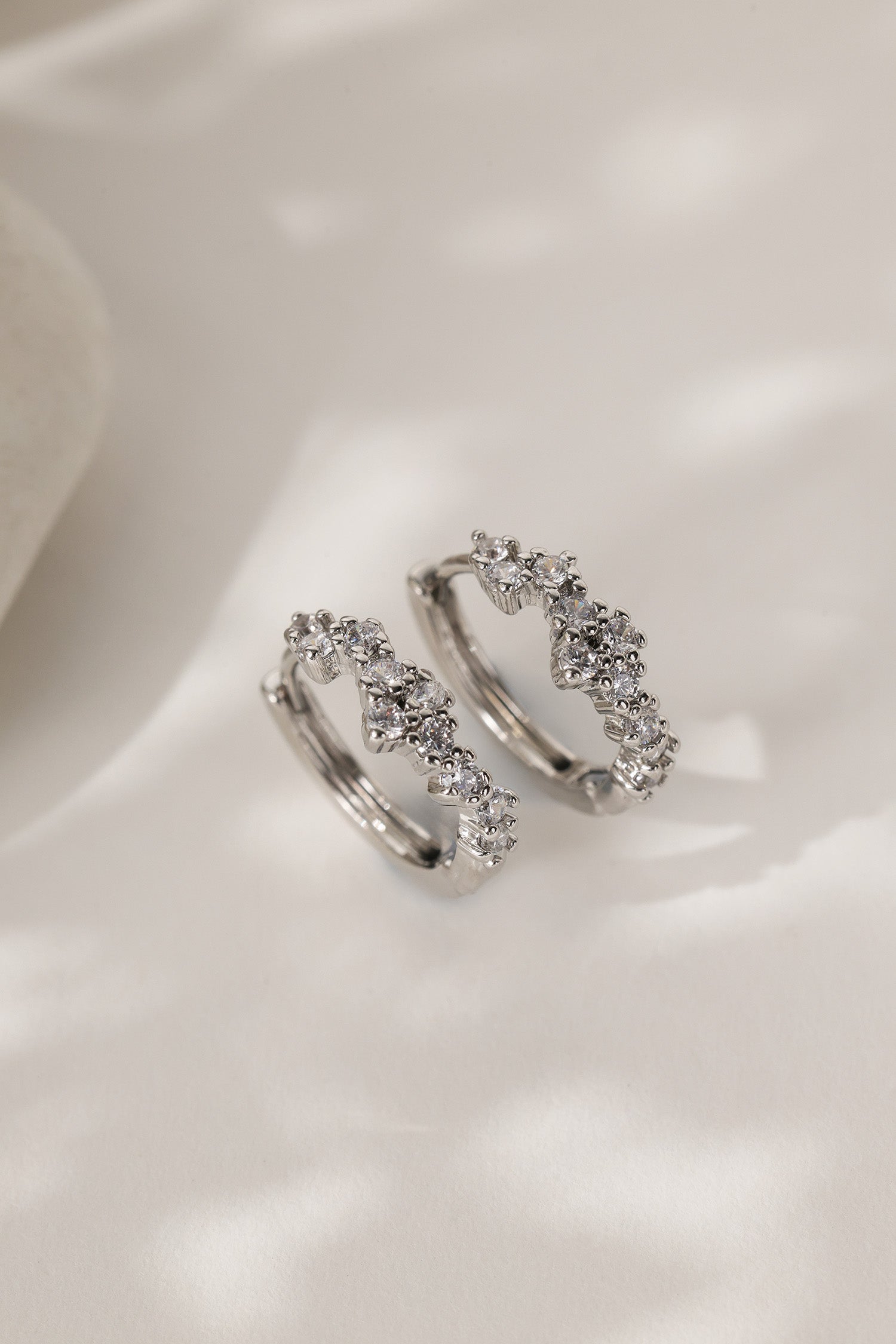 A pair of silver-toned huggie hoop earrings adorned with small clear cubic zirconia stones, displayed on a soft, light-colored fabric background.