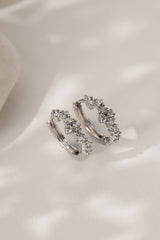 A pair of silver-toned huggie hoop earrings adorned with small clear cubic zirconia stones, displayed on a soft, light-colored fabric background.