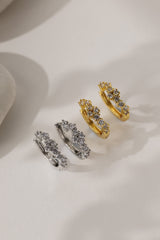 Three huggie hoop earrings with intricate floral designs, shown in silver, yellow gold, and yellow gold with clear stones, arranged on a light beige surface.