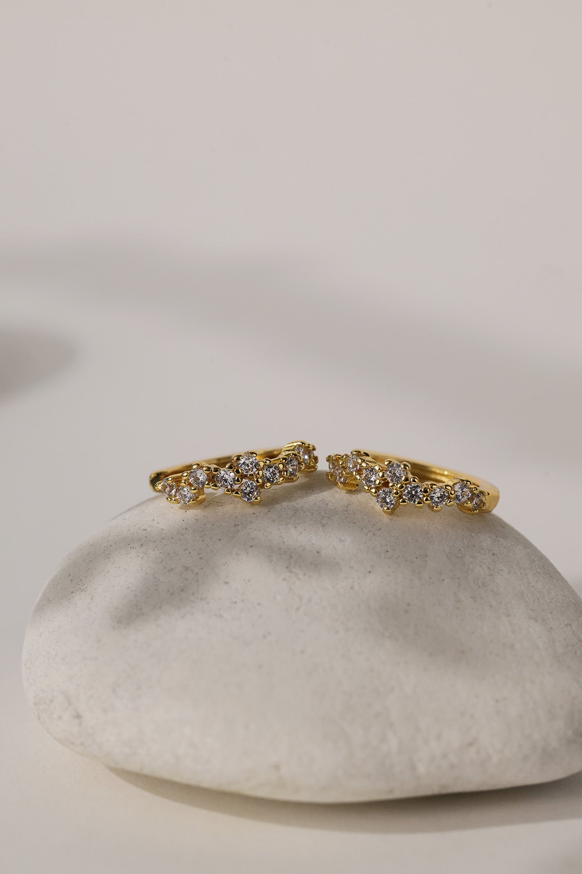 A pair of gold rings with small clear gemstones, displayed on a smooth white stone against a warm off-white background.