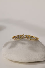 A pair of gold rings with small clear gemstones, displayed on a smooth white stone against a warm off-white background.