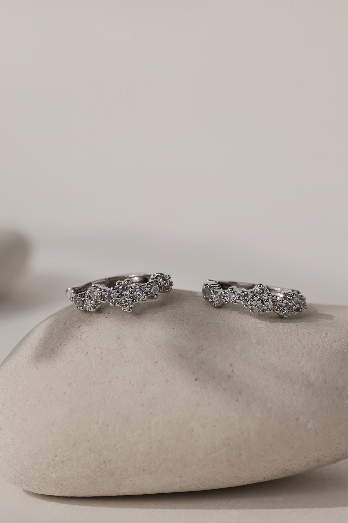 A pair of silver-toned wavy rings set with small clear stones, displayed on a smooth beige stone against a neutral light gray background.