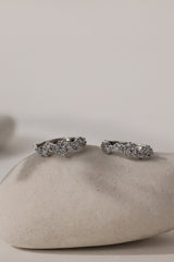 A pair of silver-toned wavy rings set with small clear stones, displayed on a smooth beige stone against a neutral light gray background.