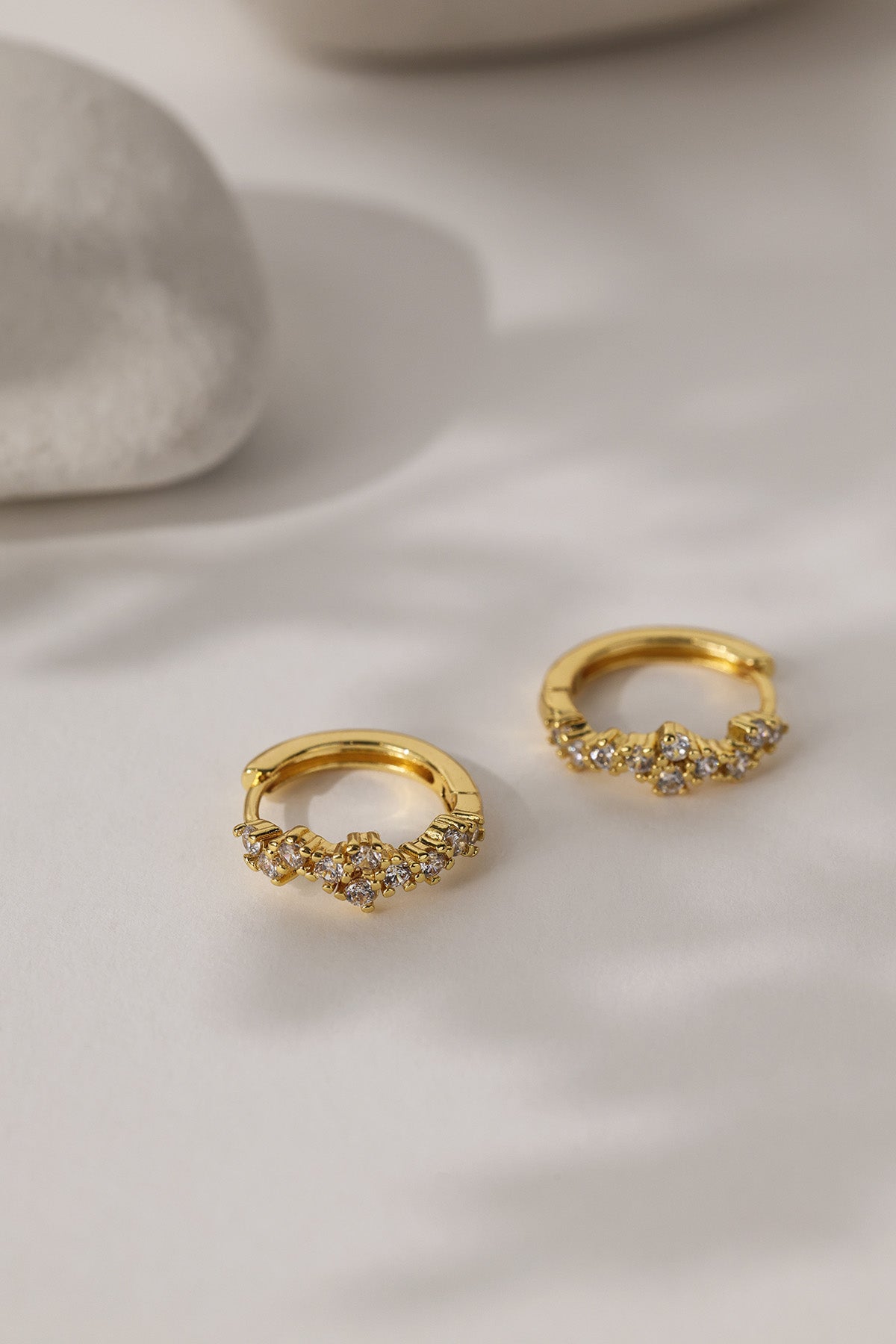 A pair of gold huggie hoop earrings adorned with small clear crystals, displayed on a soft white fabric surface with a neutral stone prop in the background.