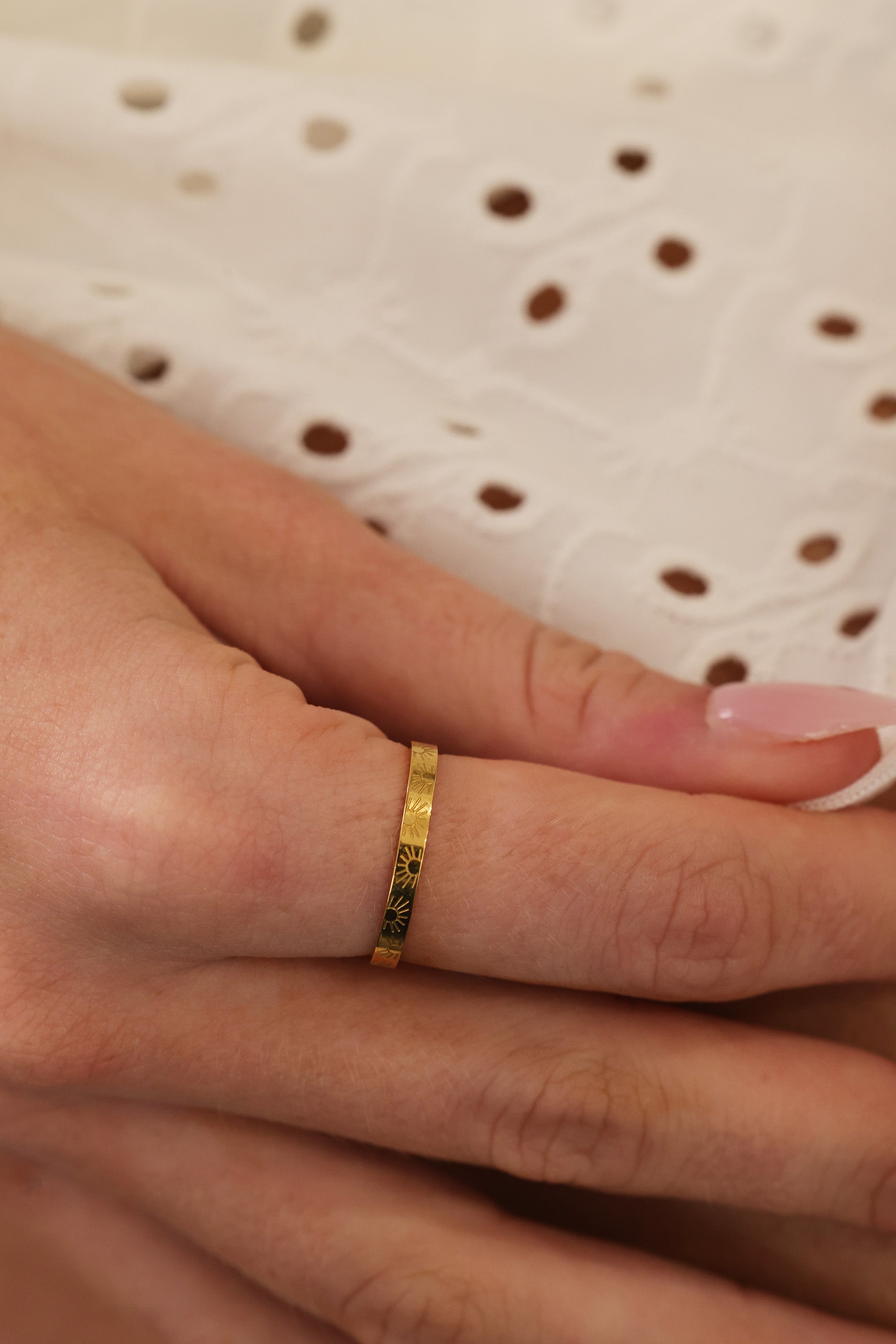 A close-up of a hand wearing a thin gold ring with small dark green stone accents, resting against a white eyelet fabric background.