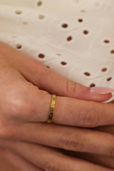 A close-up of a hand wearing a thin gold ring with small dark green stone accents, resting against a white eyelet fabric background.