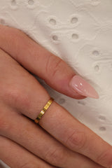 A close-up of a hand wearing a thin gold band ring with small repeating heart motifs, resting against a white textured eyelet fabric.