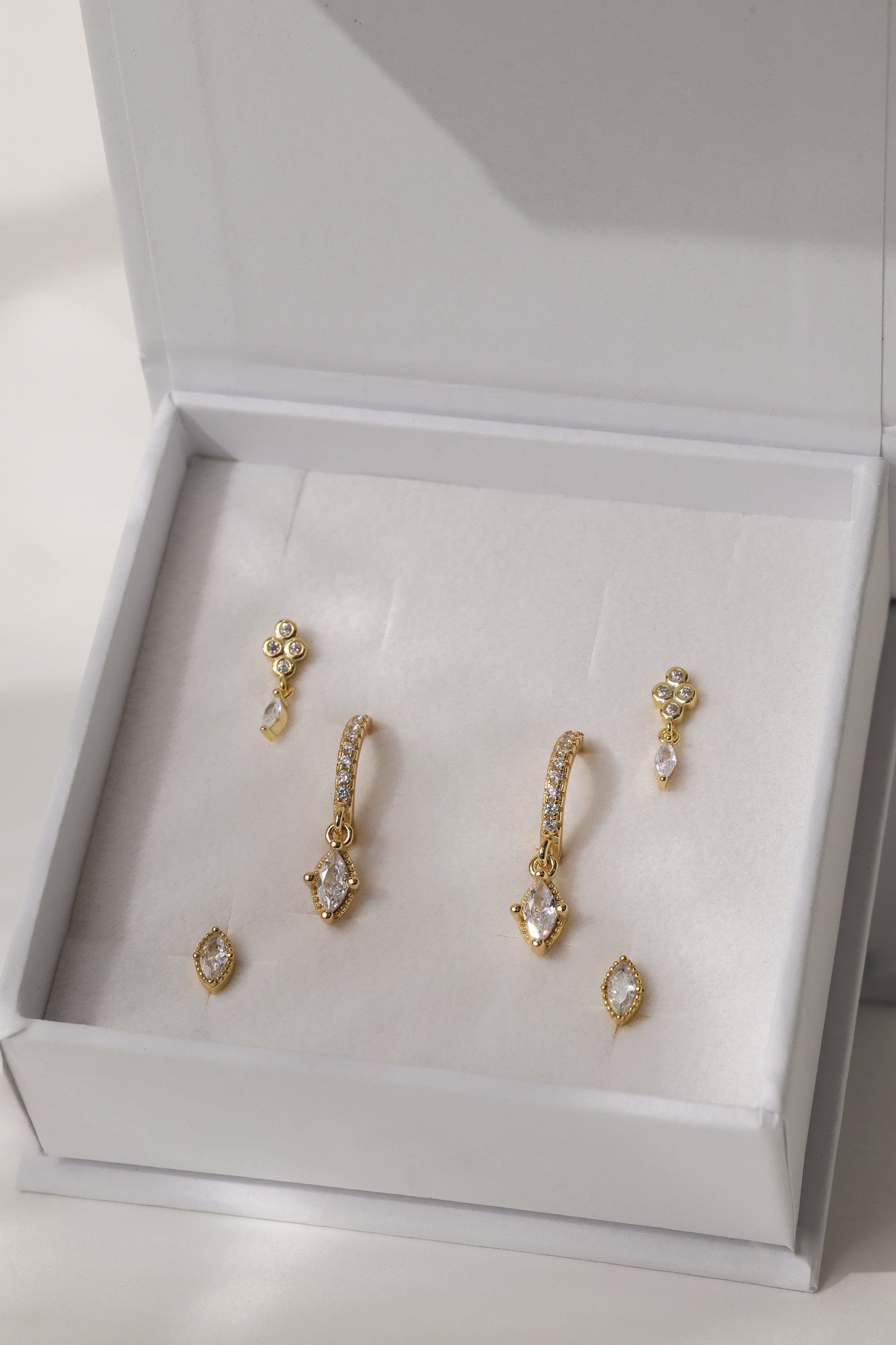 A set of gold-toned statement earrings with clear crystals, displayed inside an open white jewelry box with a cream velvet insert.