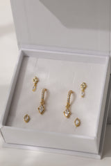 A set of gold-toned statement earrings with clear crystals, displayed inside an open white jewelry box with a cream velvet insert.