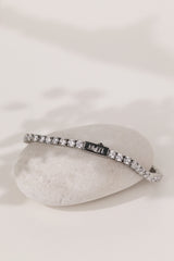 A silver tennis bracelet with a continuous row of clear gemstones, displayed on a white surface with a soft-focus white flower in the background.