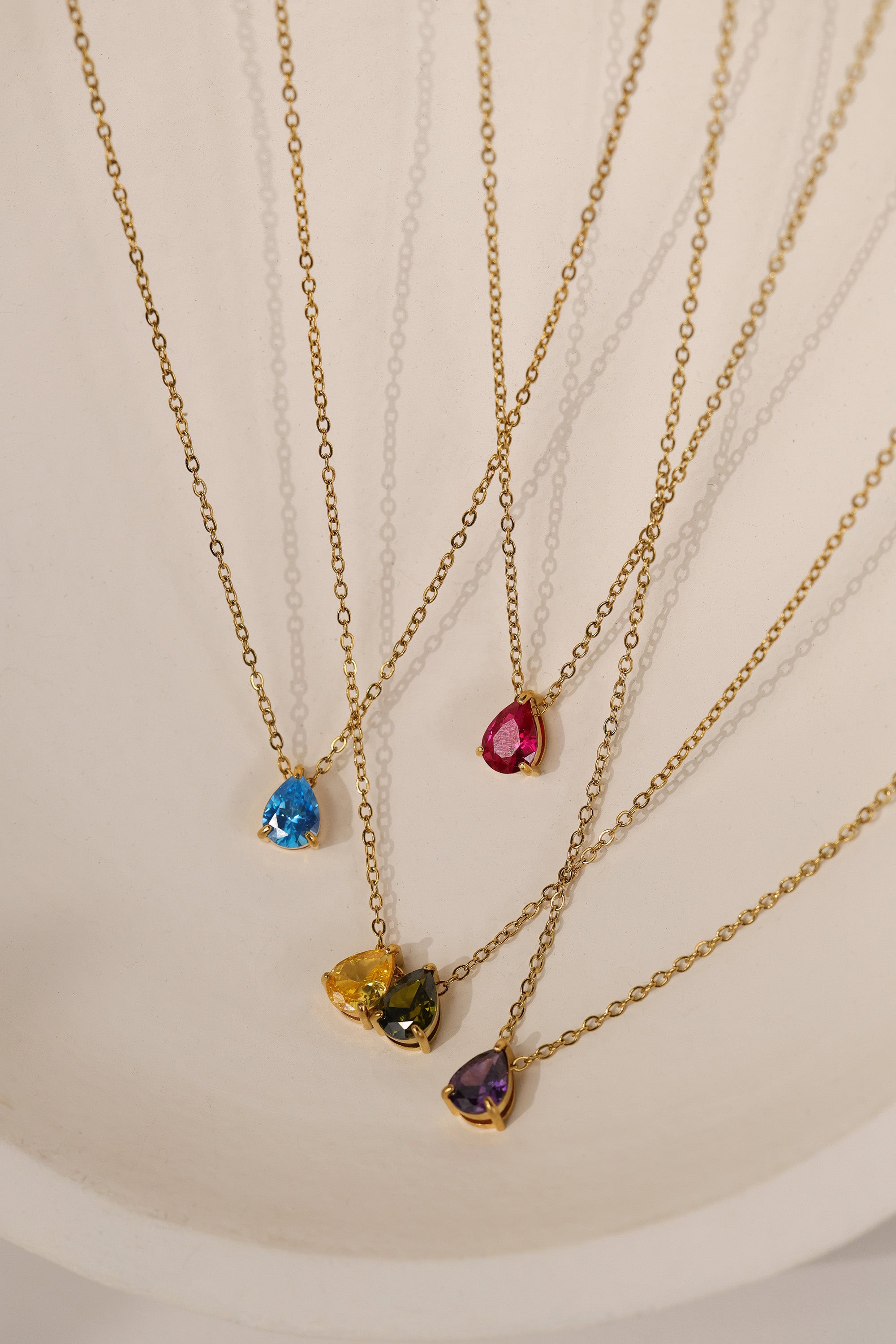 Gold birthstone necklace with two pear-cut stones on a fine cable chain, resting on a pale neutral background