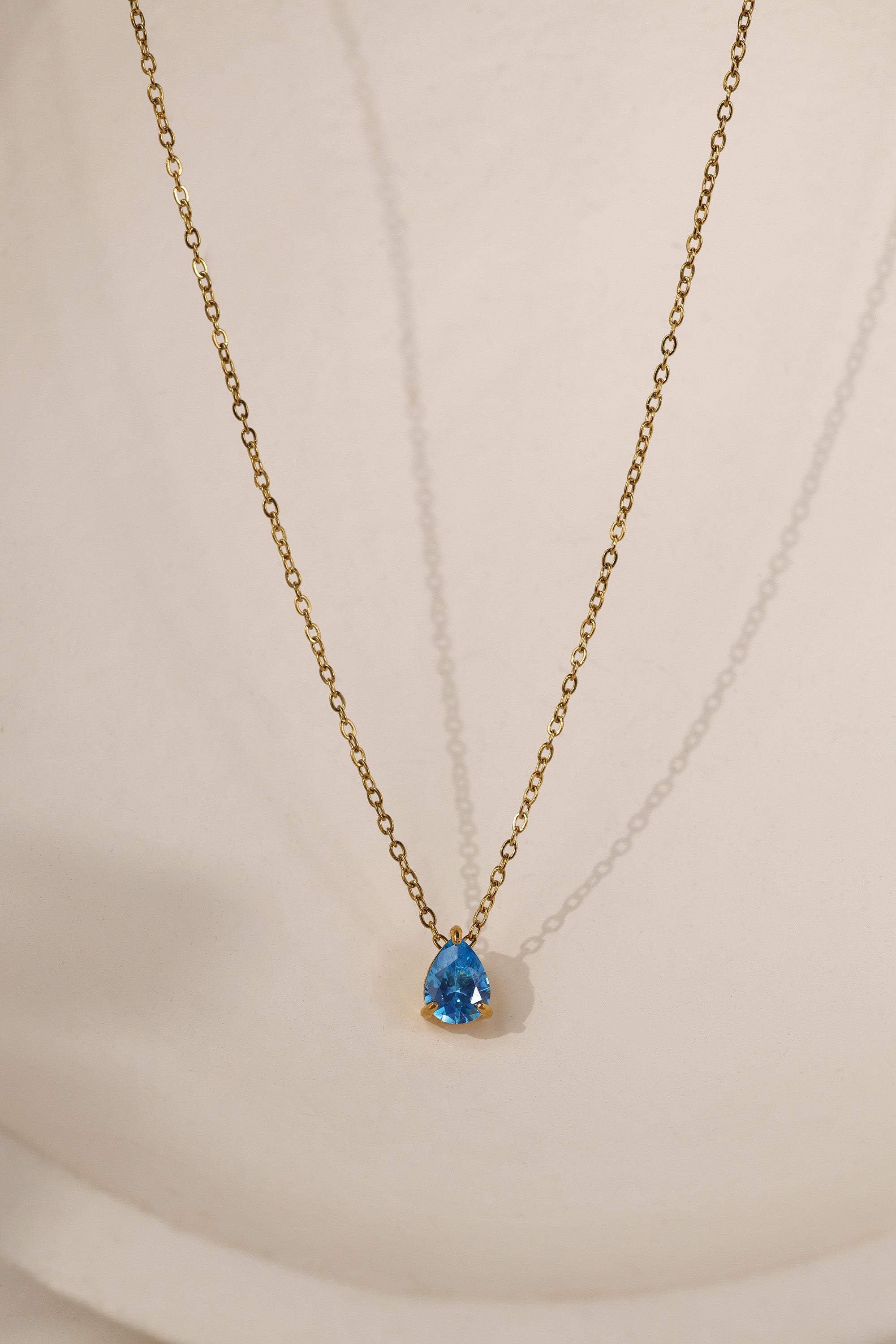 Gold birthstone necklace with two pear-cut stones on a fine cable chain, resting on a pale neutral background