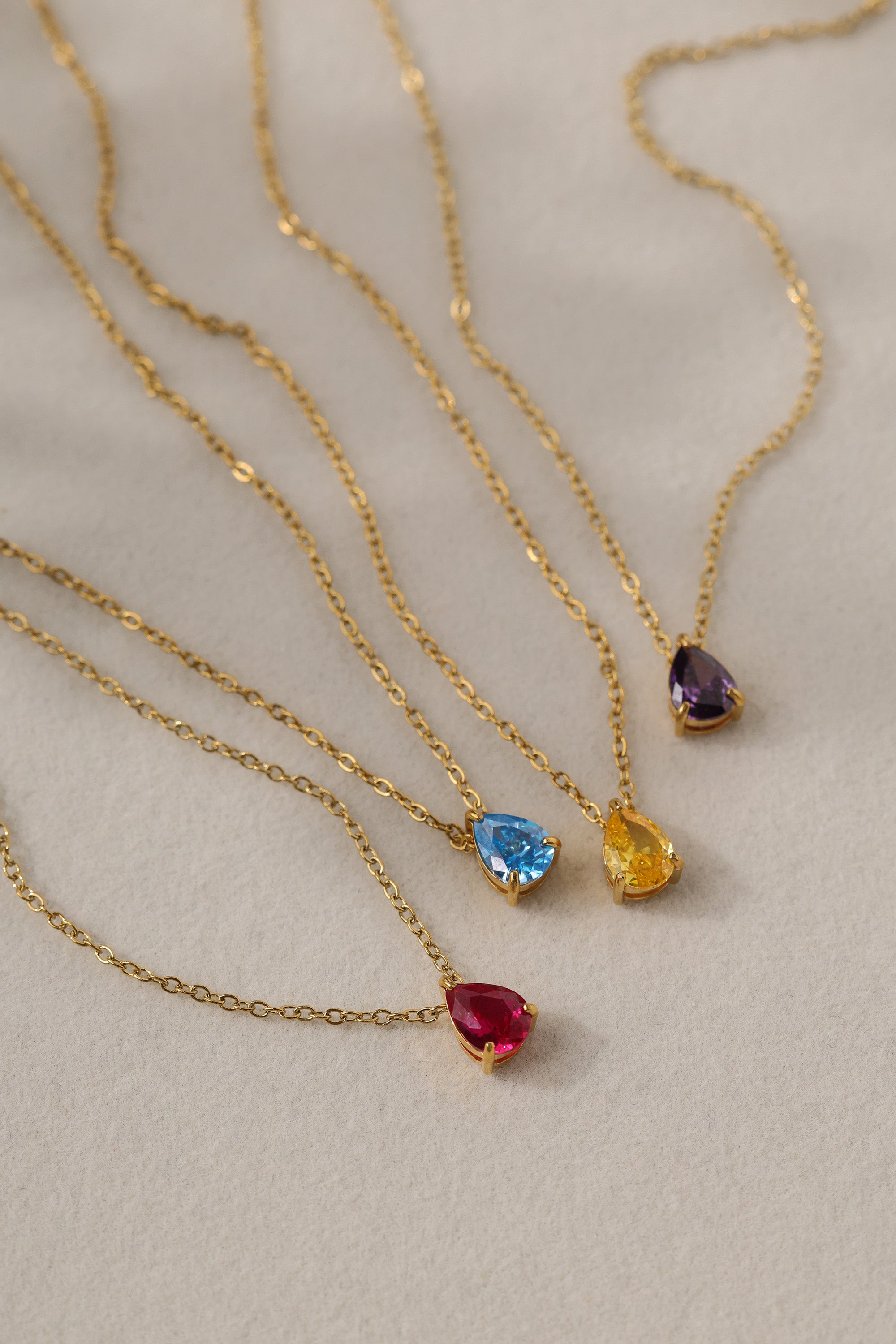 Gold birthstone necklace with two pear-cut stones on a fine cable chain, resting on a pale neutral background